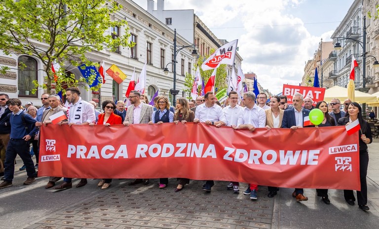 Uczestnicy pochodu pierwszomajowego niosący czerwony baner z hasłem „Praca Rodzina Zdrowie” w centrum miasta.